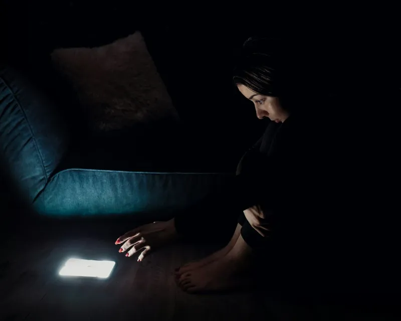 A woman sitting in the dark reaching for her phone. (Photo Credit Inspa Makers on Unsplash) Addictions header image