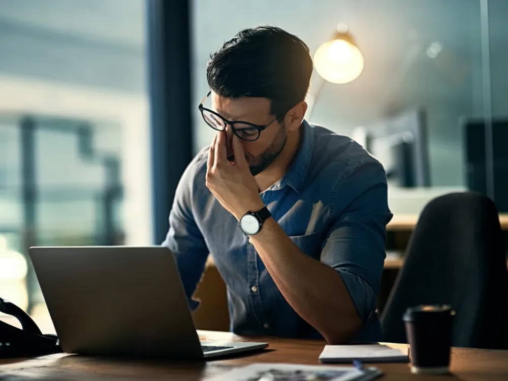 A man sitting at a desk with his laptop looking exhausted